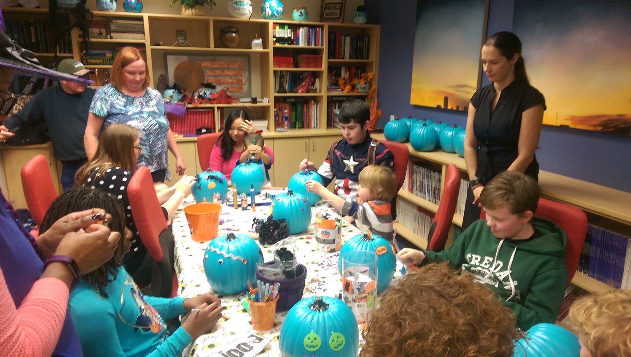 Children and teachers sitting around a table covered in halloween decorations. The children are decorating pumpkins that have been painted teal