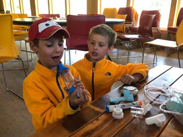 Two young boys in a classroom setting, sitting at a table with pieces of asthma-related medical equipment. They are holding one of the pieces and learning how it is used.
